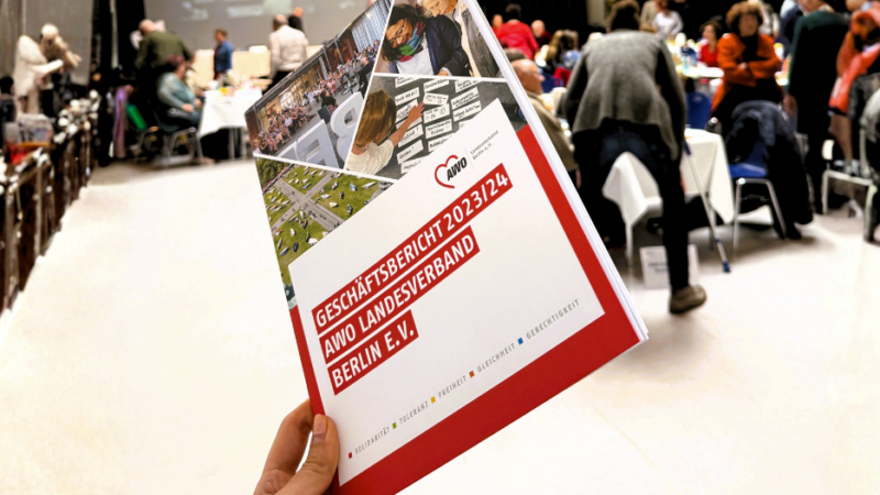 Eine Hand hebt den Geschäftsbericht 2023–2024 in die Höhe, während im Hintergrund die lebhafte Atmosphäre der Landeskonferenz zu sehen ist.
