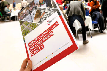 Eine Hand hebt den Geschäftsbericht 2023–2024 in die Höhe, während im Hintergrund die lebhafte Atmosphäre der Landeskonferenz zu sehen ist.