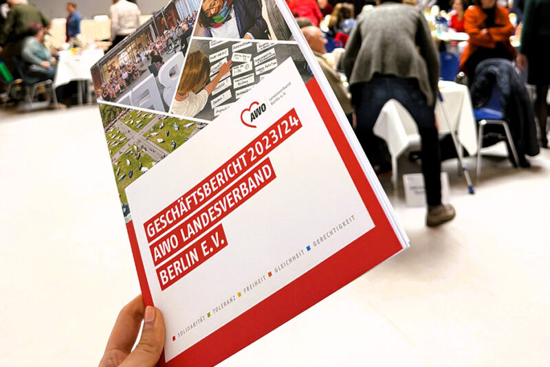 Eine Hand hebt den Geschäftsbericht 2023–2024 in die Höhe, während im Hintergrund die lebhafte Atmosphäre der Landeskonferenz zu sehen ist.